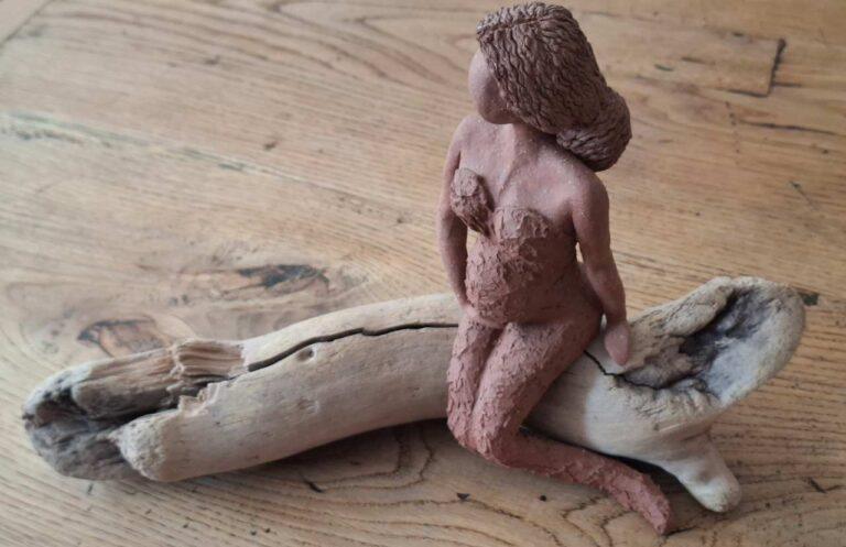 Weibliche Tonfigur die auf einem Treibholzstück sitzt.