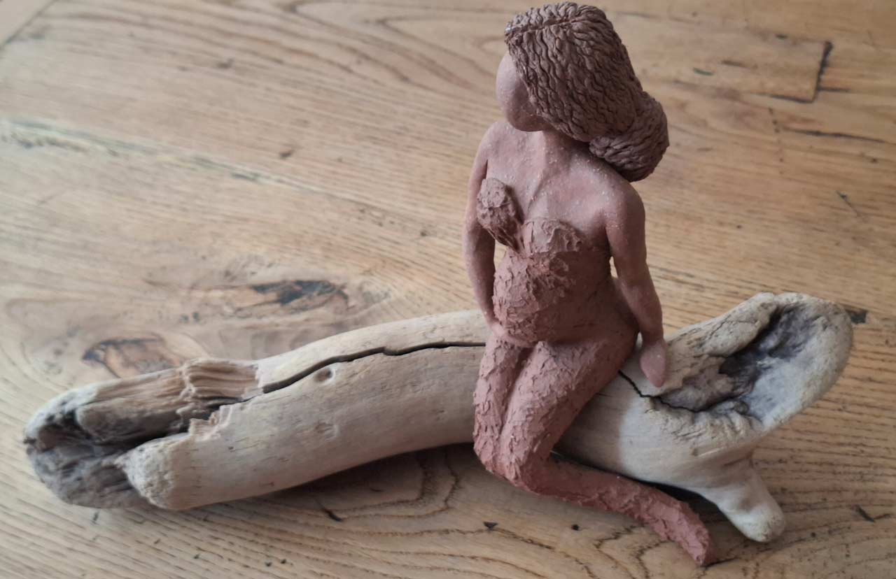 Weibliche Tonfigur die auf einem Treibholzstück sitzt.